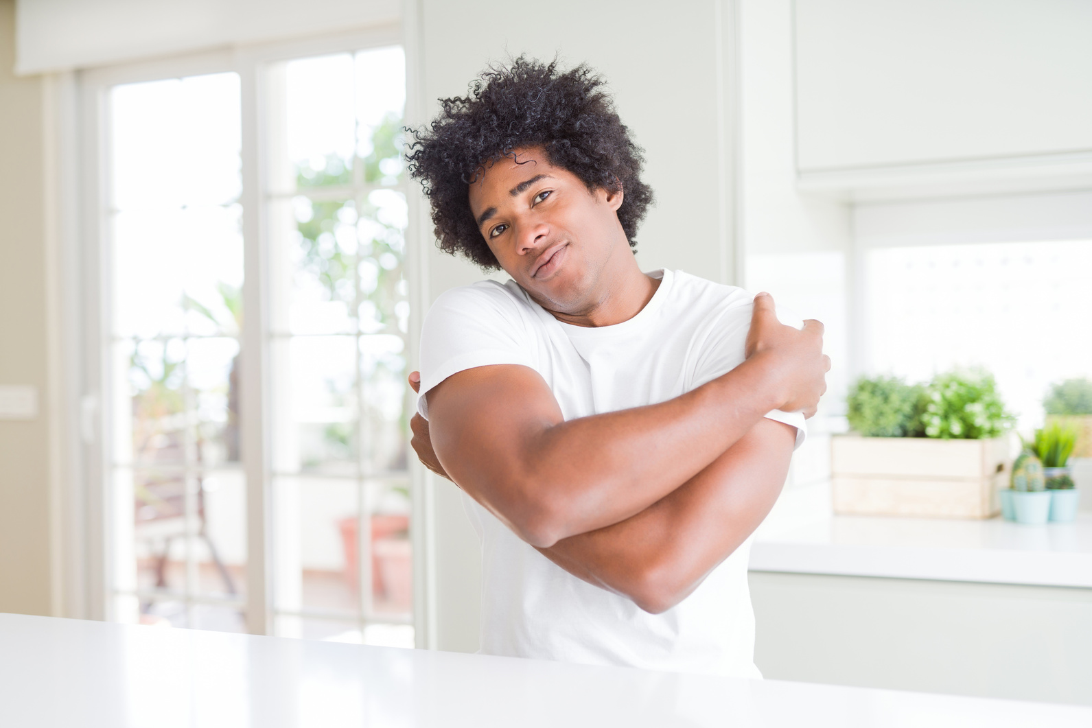 Young african american man wearing casual white t-shirt sitting at home Hugging oneself happy and positive, smiling confident. Self love and self care